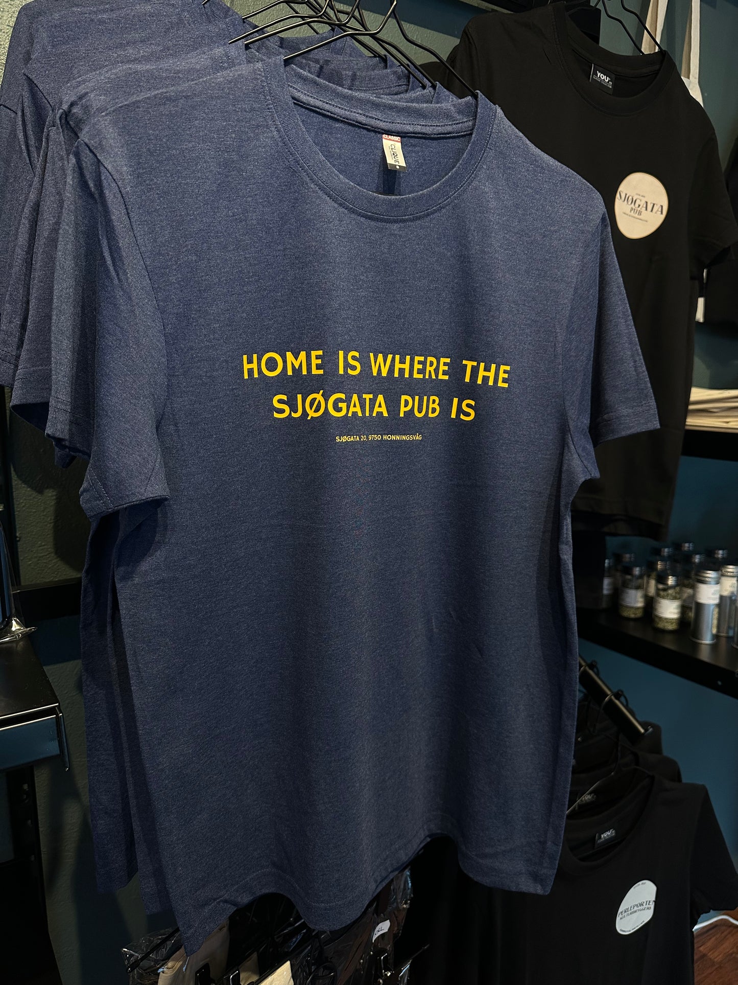 Mørk t-skjorte med gul tekst HOME IS WHERE THE SJØGATA PUB IS på kleshenger