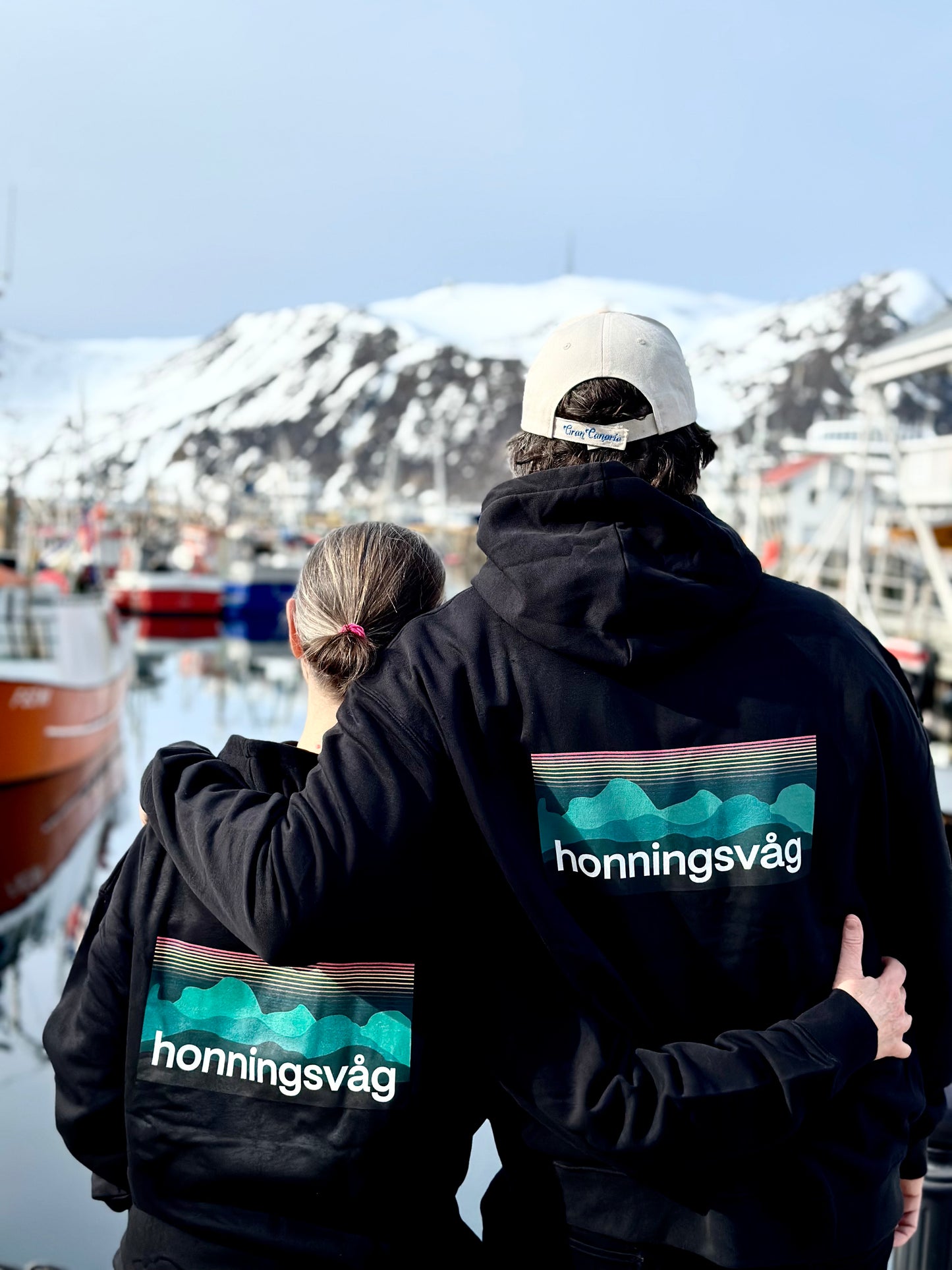 To personer bakfra i svarte hettegensere med honningsvåg-trykk på ryggen, ved havna