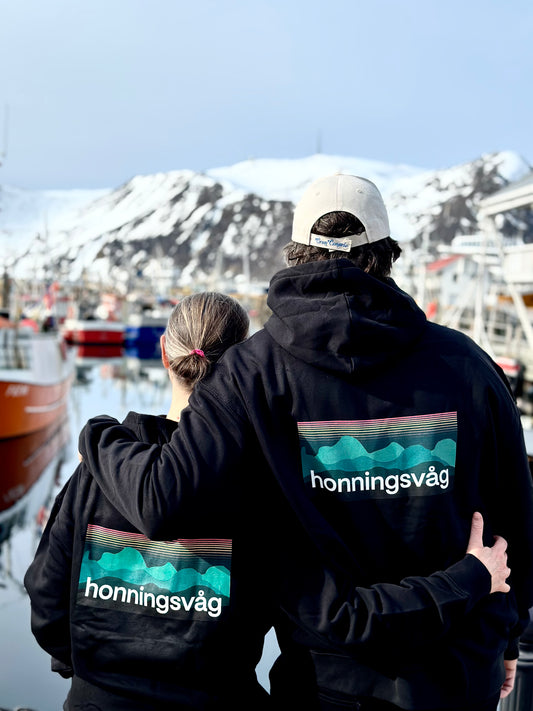 To personer bakfra i svarte hettegensere med honningsvåg-trykk på ryggen, ved havna