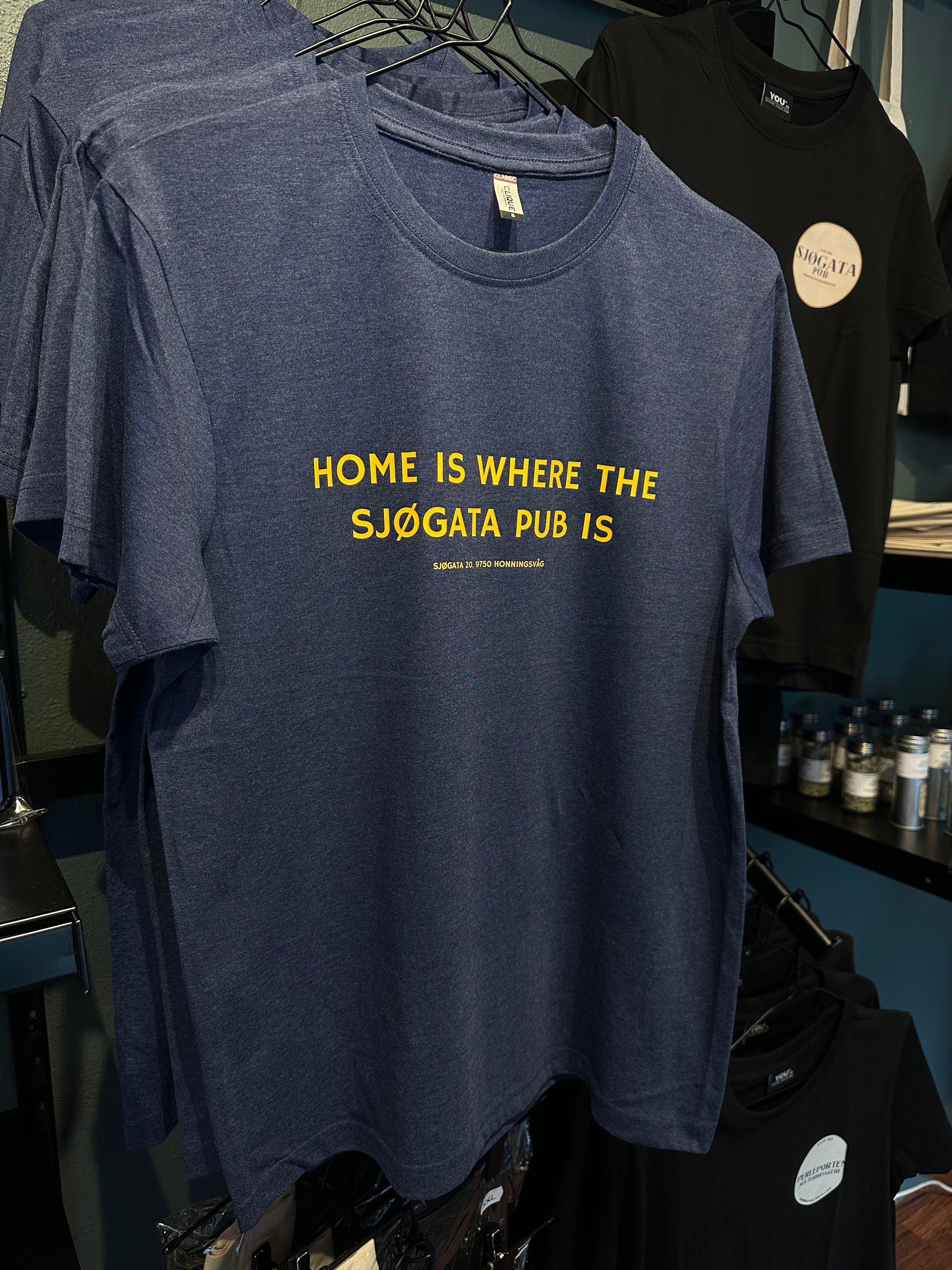 Mørk t-skjorte med gul tekst HOME IS WHERE THE SJØGATA PUB IS på kleshenger